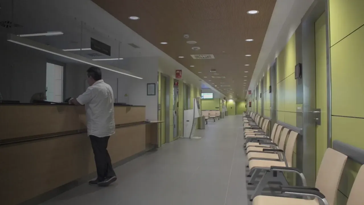 Pasillo hospital con recepción, personal, sillas a la derecha y paredes verdes bajo luz