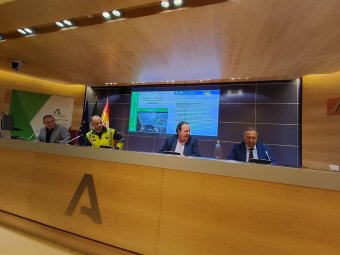 Quatre homes a taula de conferència, amb pantalla i logos de fons.