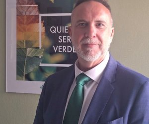 Hombre de aspecto profesional con traje azul oscuro y corbata verde, frente a una decoración botánica, depuestamente en un espacio de oficina.