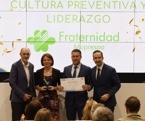 Cuatro personas en un escenario, dos hombres sostienen un certificado, fondo con texto y logo.