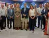 Grupo de personas de distintasedades y géneros, de pie en fila, en interior con fondo azul y luces.