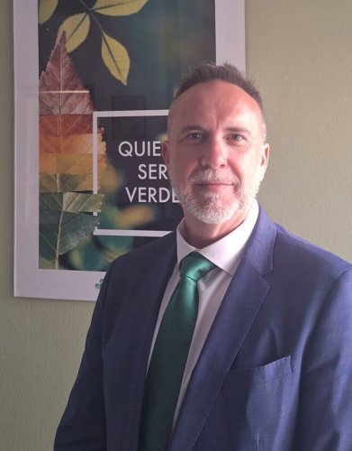 Hombre de aspecto profesional con traje azul oscuro y corbata verde, frente a una decoración botánica, depuestamente en un espacio de oficina.